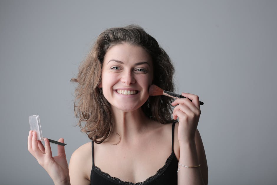 A joyful woman applies makeup with a brush, posing in a studio with a bright smile.