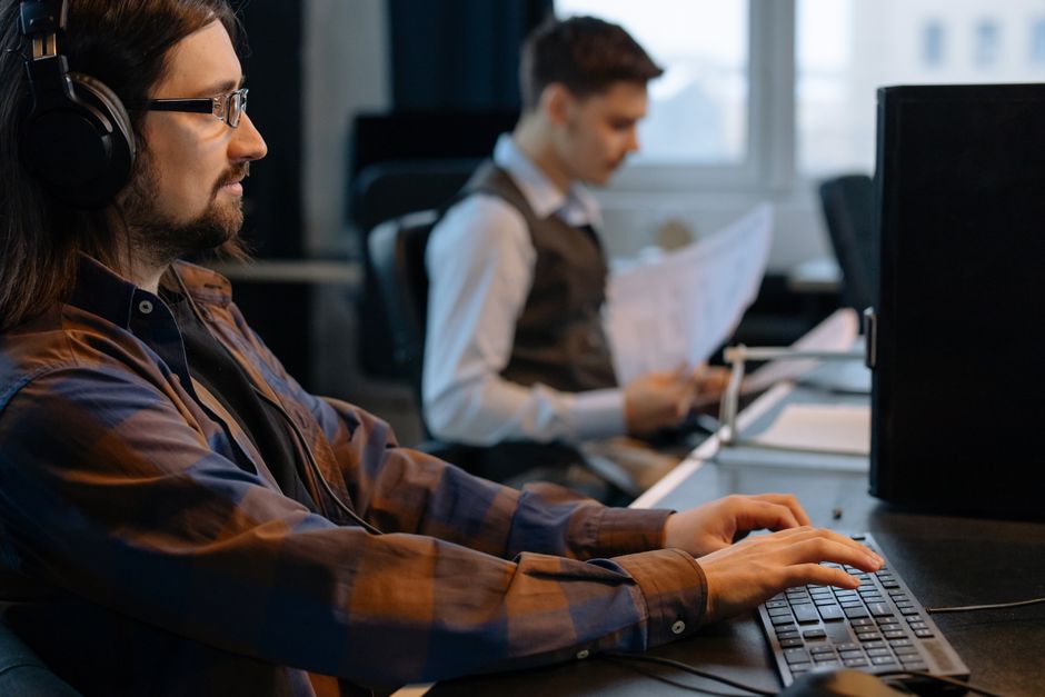 Bearded male developer in headphones coding on a laptop in a modern office setting.